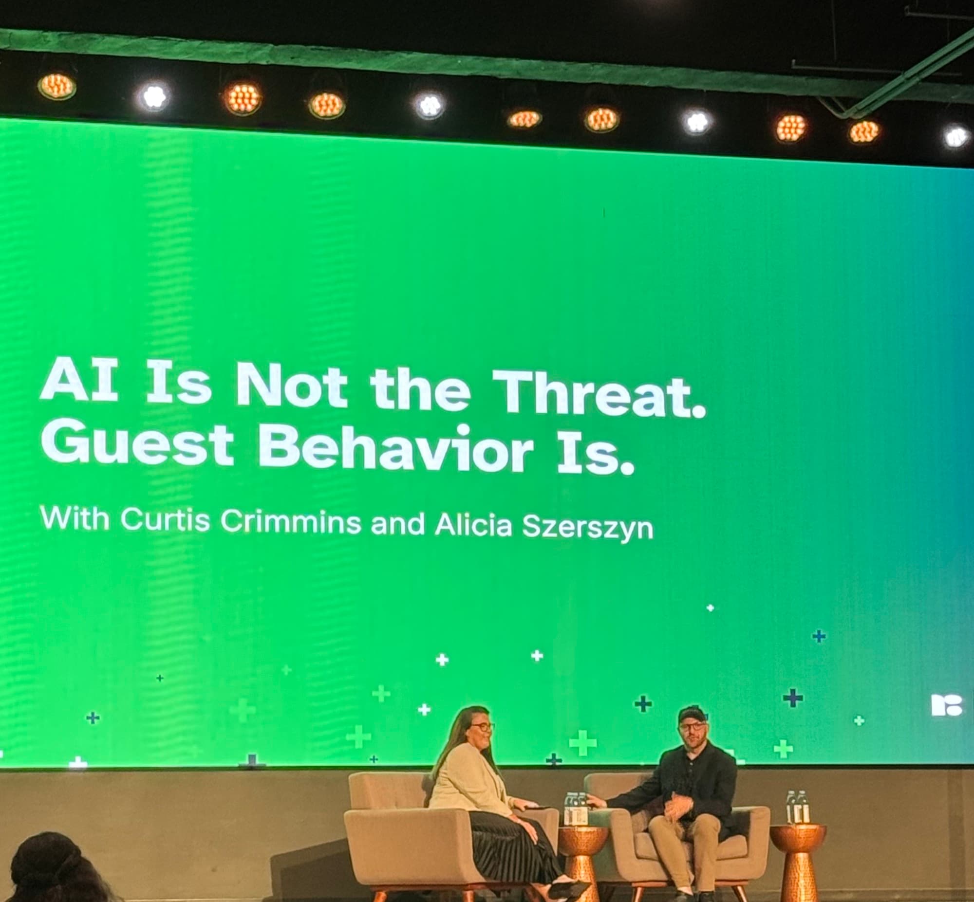 Curtis on a conference stage in conversation beneath a screen reading AI Is Not the Threat. Guest Behavior Is.
